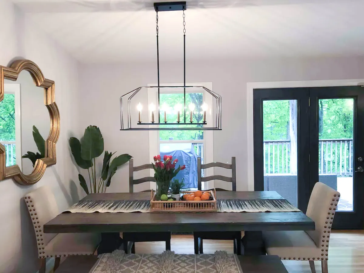 A warmly lit dining area with a central table ready for gatherings, highlighted by a chandelier, fresh flowers, and elegant decor. Doors in the scene suggest a connection to other spaces and possibly an easy transition to outdoor or communal activities.