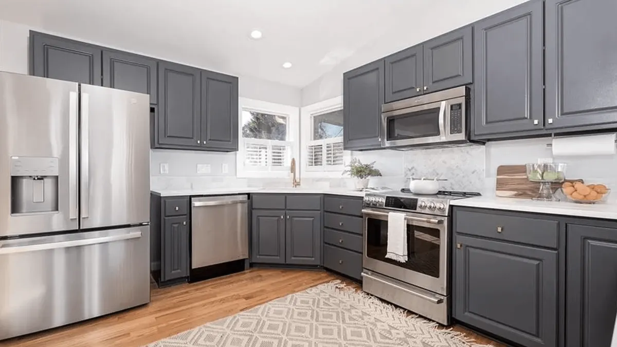 A modern kitchen featuring new stainless steel appliances, sleek gray cabinets, a bright white countertop, and a woven rug underfoot. The space is designed for both cooking and gathering, with ample workspace and a welcoming mood ideal for entertaining.