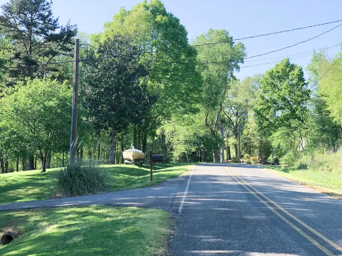 The photo shows a tranquil roadside scene near the house, with a view down a tree-lined road, a boat, a mailbox, and peaceful greenery—suggesting easy access to outdoor activities like walking, biking, or fishing. The setting sun adds a golden, welcoming glow.