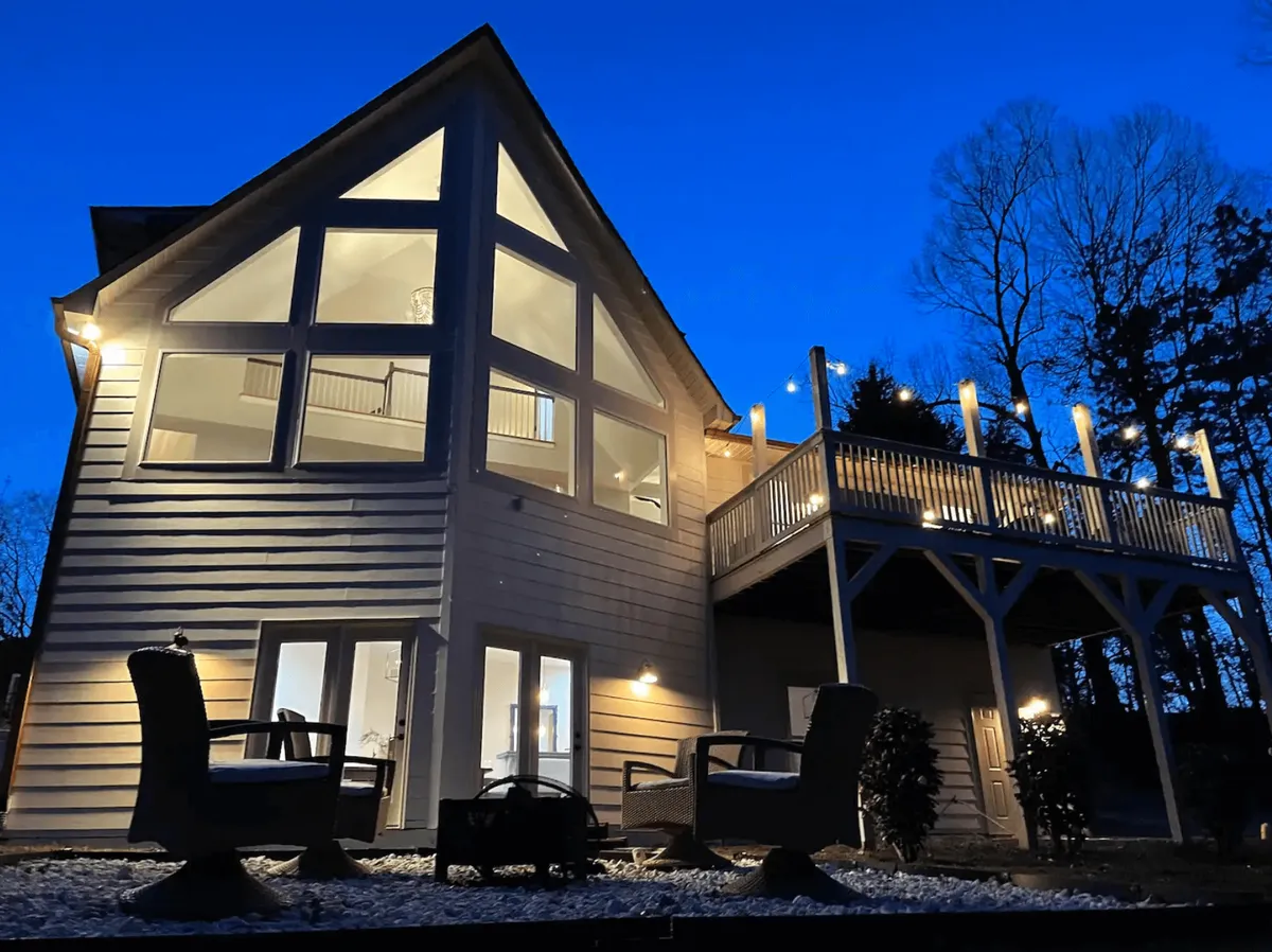 The hero image showcases a stunning lake house exterior, featuring expansive windows that invite natural light, a spacious deck adorned with string lights, and inviting outdoor seating perfectly arranged for dining or cozy evenings under the stars. The serene lakeside ambiance is enhanced by the tranquil surroundings and the promise of memorable moments spent outdoors.
