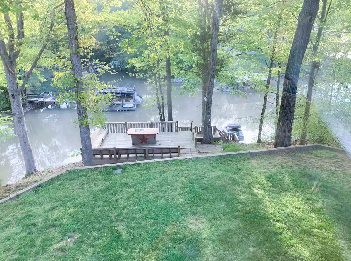 A spacious deck area overlooking the serene lake, featuring a table perfect for outdoor dining. The view includes calm water, lush trees surrounding the property, and hints of fishing or boating activities ready to be enjoyed from the dock.