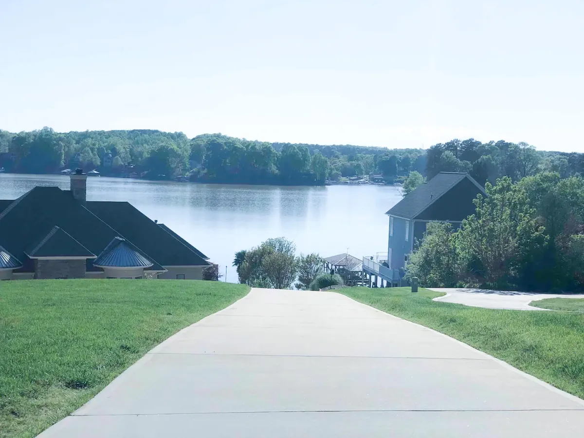 A tranquil scene of the lake, with a meandering pathway lined by greenery and nearby homes. The perspective invites viewers to imagine strolling along the shore, enjoying the peaceful surroundings, and seeing boats out on the water.