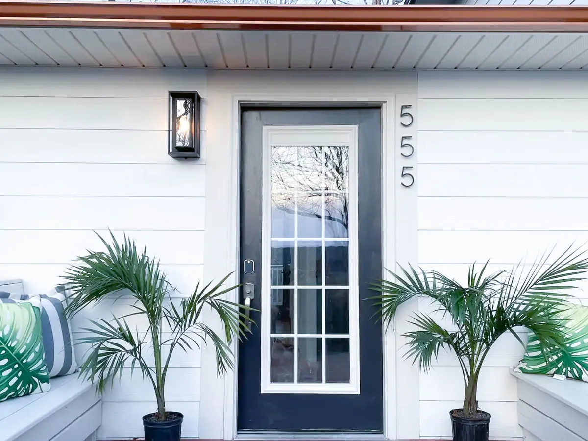 The image shows the welcoming front entry of the home, featuring the front door framed by cheerful potted plants and decorative cushions on the porch. The house number is clearly visible, and the setting is inviting, suggesting a place where guests are greeted and families gather before entering the home.