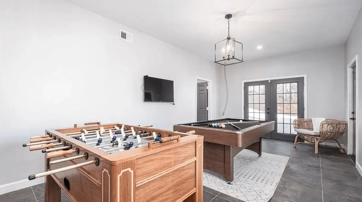 A spacious and lively game room featuring a foosball table and pool table, set beside large windows that flood the area with natural light. The scene includes a wicker chair for casual seating, and the open floor plan encourages social interaction and recreation.