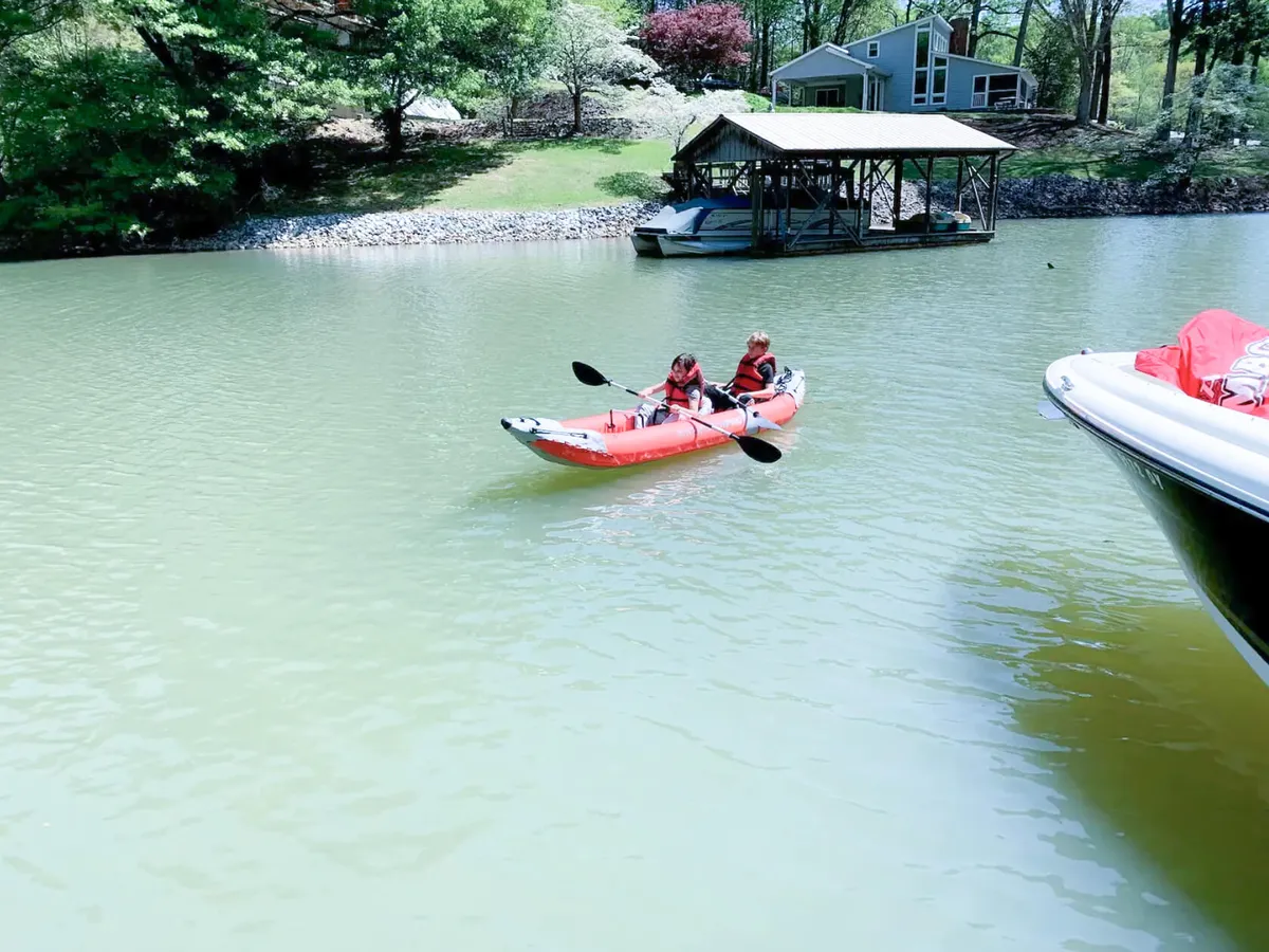 A red kayak glides past a private dock surrounded by lush greenery, with the lake reflecting calm skies. The image conveys adventure and relaxation as someone paddles on the lake, highlighting personal watercraft access and the natural beauty of the water’s edge.
