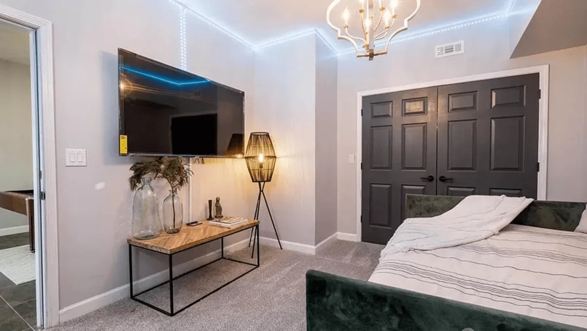 A stylish bedroom with a green velvet bed, modern chandelier, wood accents, and a flat-screen TV. Decorative lamps and a wooden table with vases add warmth and character, while the cozy layout encourages relaxation or leisure time.