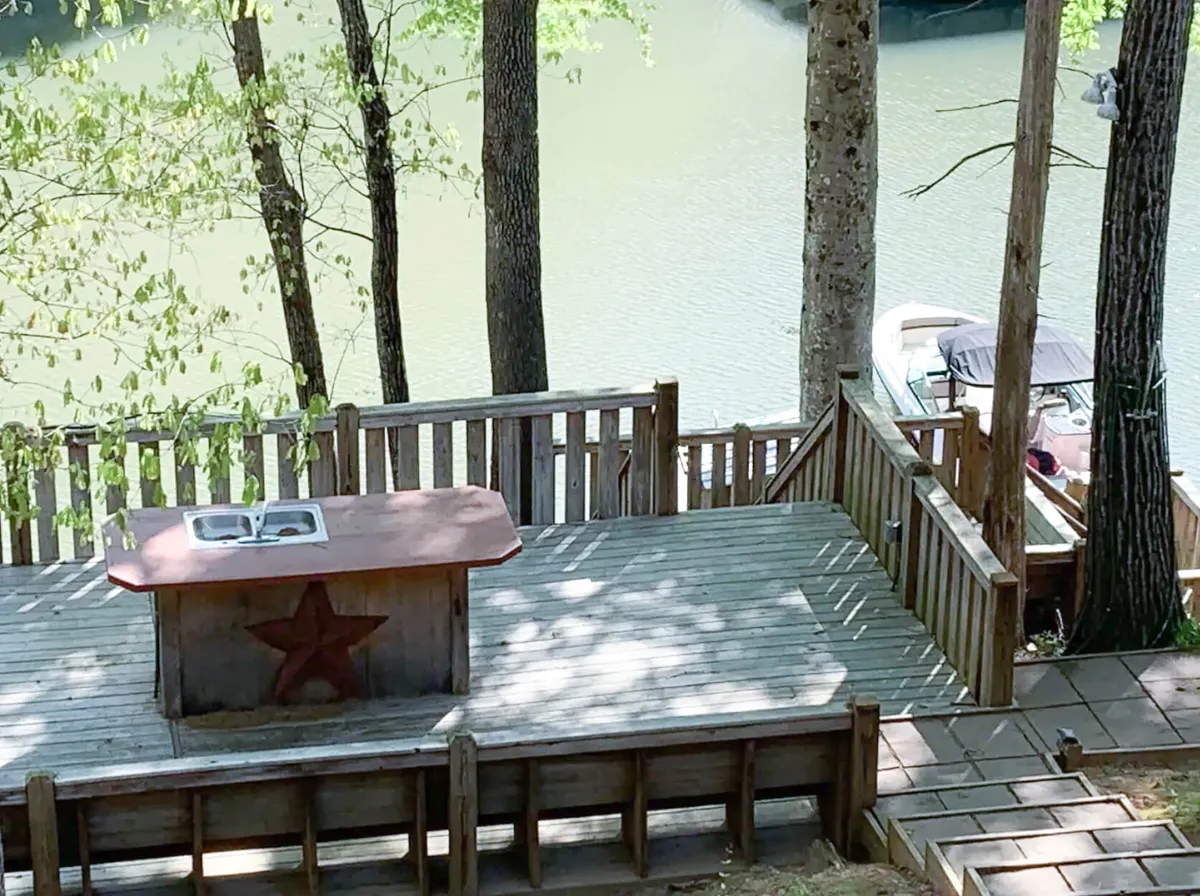 A stylish outdoor deck faces the water, featuring a built-in Charbroil grill and sink with a star design, surrounded by tall trees. The deck overlooks a serene lake with a boat moored nearby, hinting at direct water access and opportunities for fishing or boating.